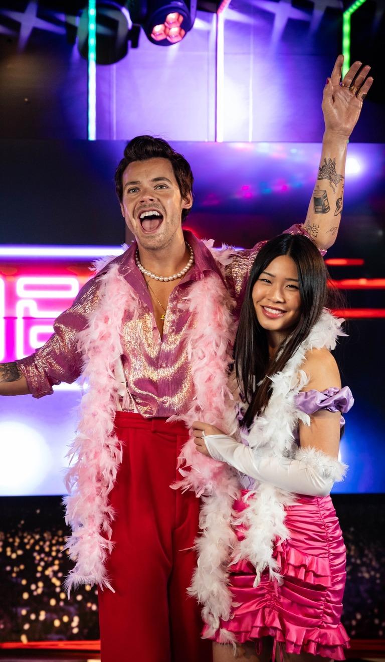 Women is wearing a pink dress with a white feather bower stood next to a model figure of Harry Styles which is dressed in a red and pink outfit.