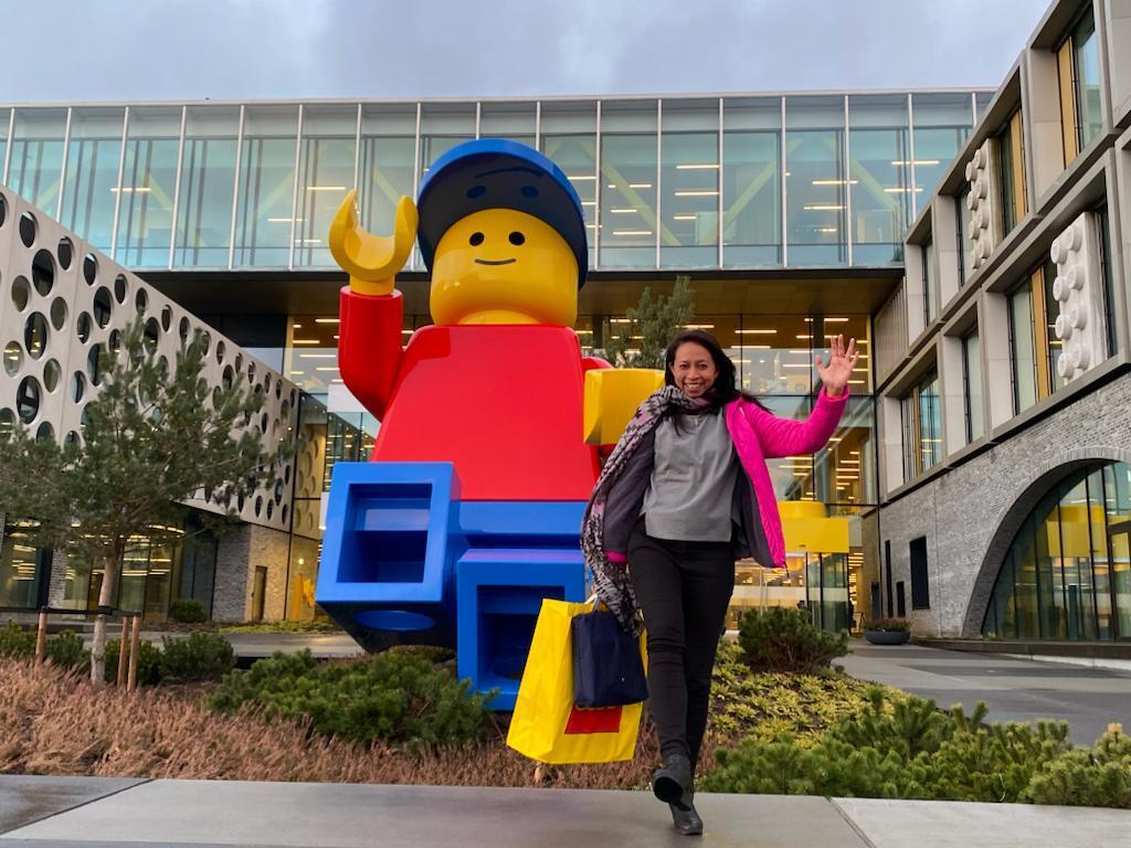 Women is wearing a grey jumper and a bright pink coat and is jumping in the air outside of a building with a giant LEGO® figure behind her.