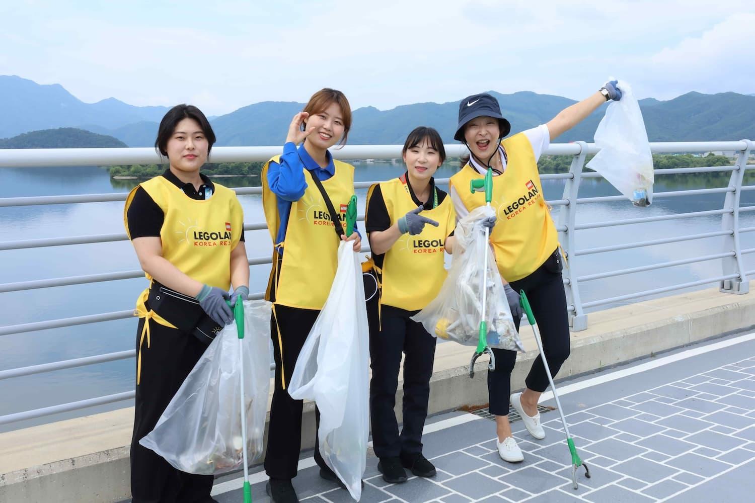 4 female LEGOLAND employees in yellow vests pose with litter pickers and bin bags. Behind them is a body of water with scenic hills.