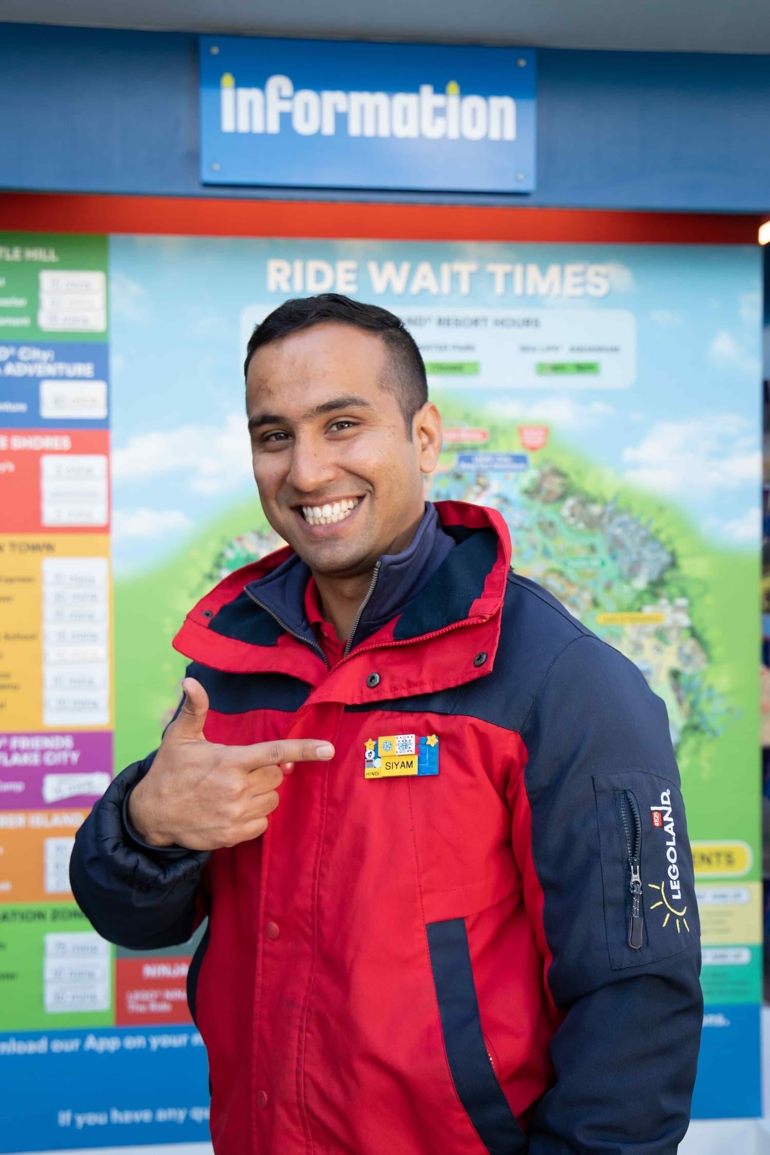 Male LEGOLAND worker in a red and navy LEGOLAND branded coat is standing in front of a noticeboard. He smiles and points at his name badge.