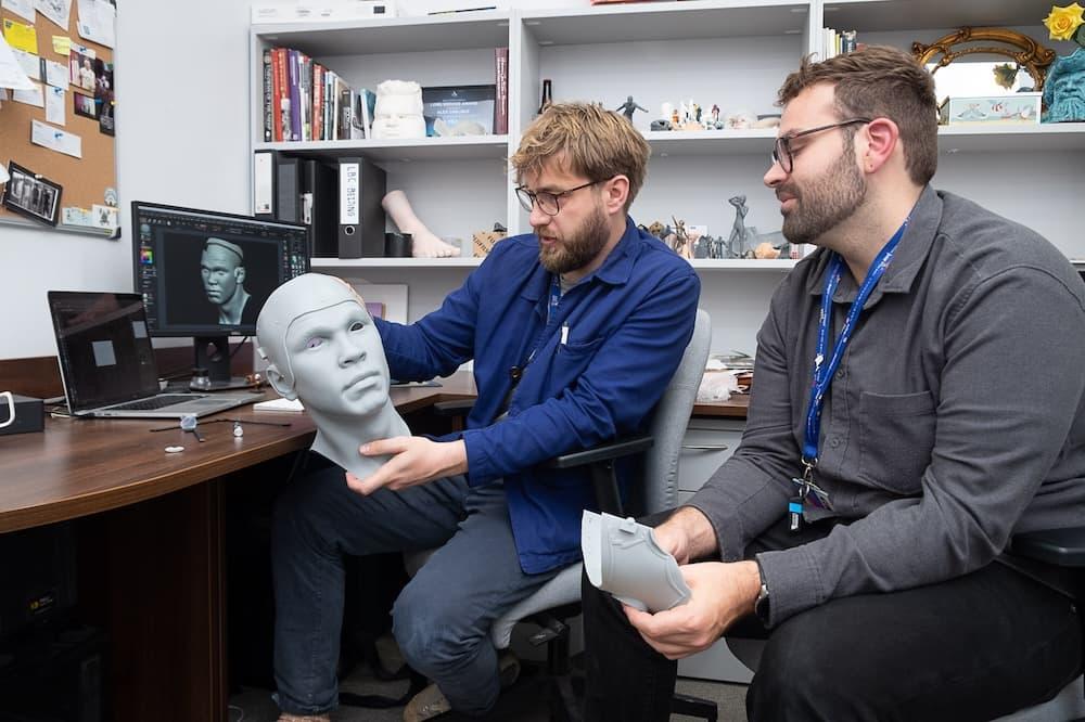 In an office, a man is sitting down holding a 3D model of a head. He shows another man who holds another part of the model.