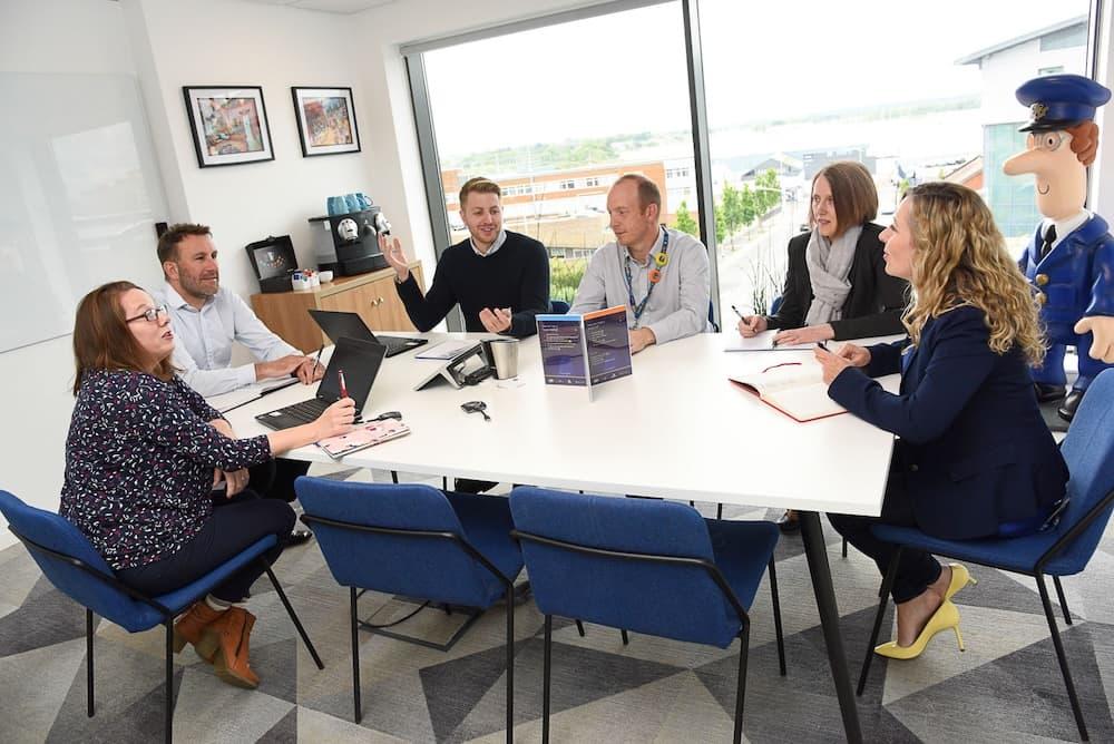 6 coworkers sit around a table in an office in front of a window in conversation. There is a postman pat statue in the corner.