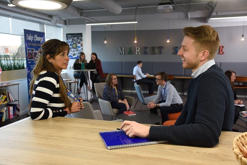 In a populated seating area a man sat with a blue notebook smiles towards a female in a black and white striped jumper.