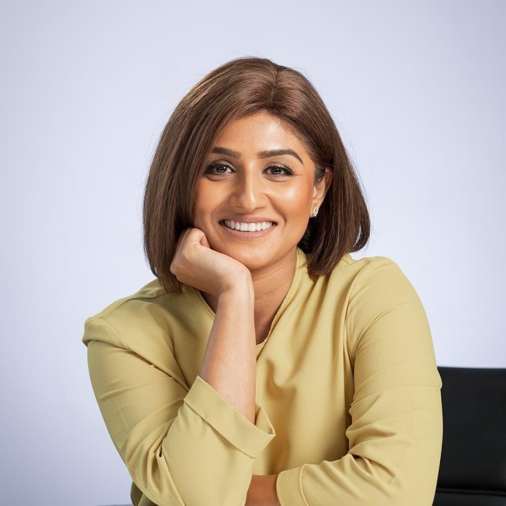 A headshot of a women staring at the camera. She is smiling and has her right hand rested against her chin. She is wearing an olive green shirt.