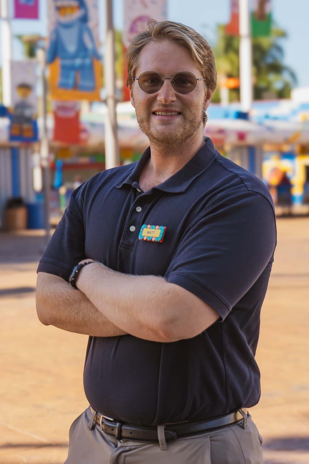 A headshot of Matt, an assistant manager. He smiles into the camera with his arms folded, sunglasses on and navy polo tucked in.