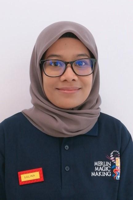Women is wearing a navy blue polo shirt with the Merlin Magic Making logo on and a beige headscarf and glasses.