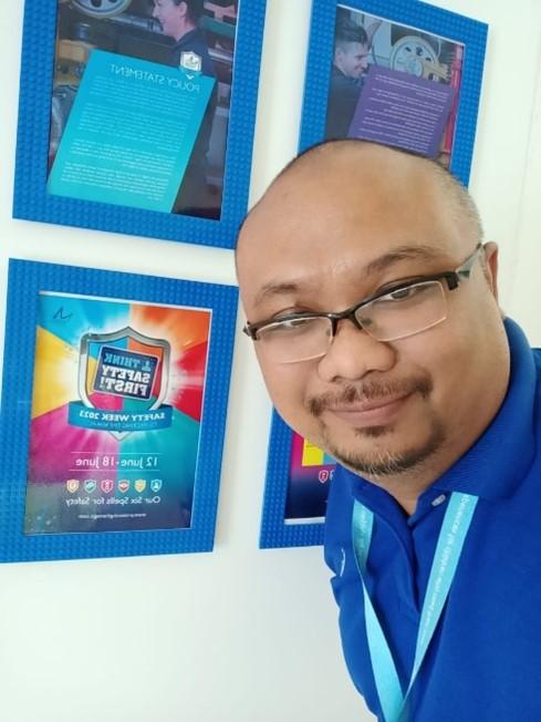 Bald man wearing a blue polo shirt and lanyard and black frame glasses is posing in front of four blue posters stuck on a white wall.