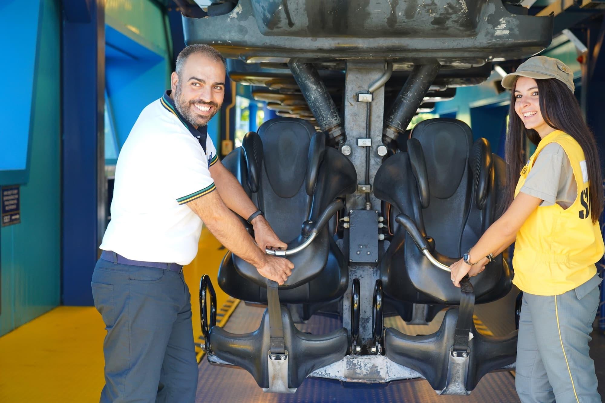 Women wearing a yellow Gardaland top and man in a white polo shirt and stood in front of a rollercoaster adjusting the seat and security mechanism.