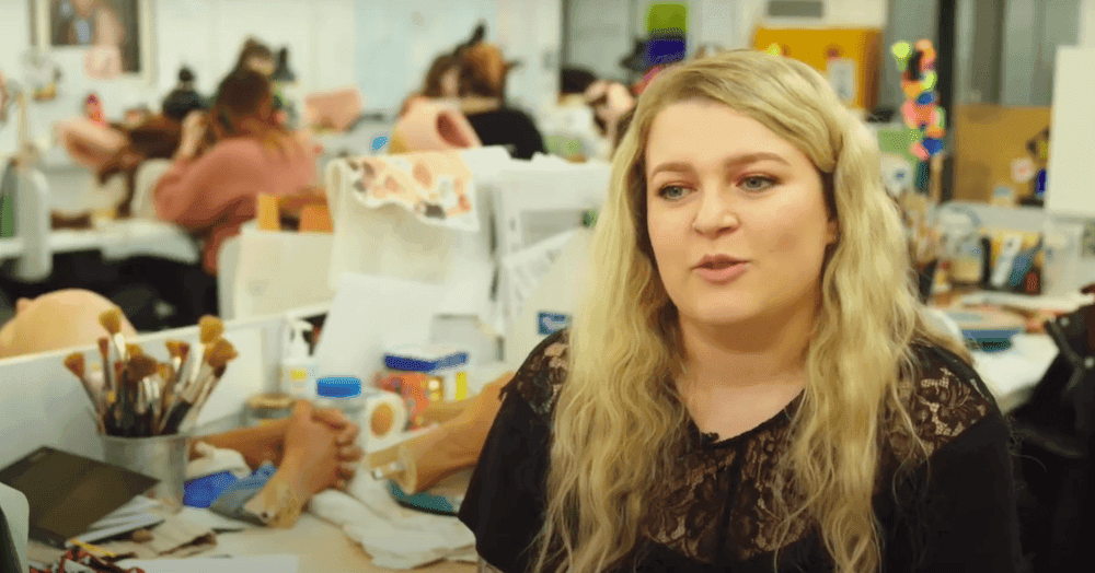 Women with long blonde hair in a black outfit is sat in an art studio with paintbrushes and model figures.