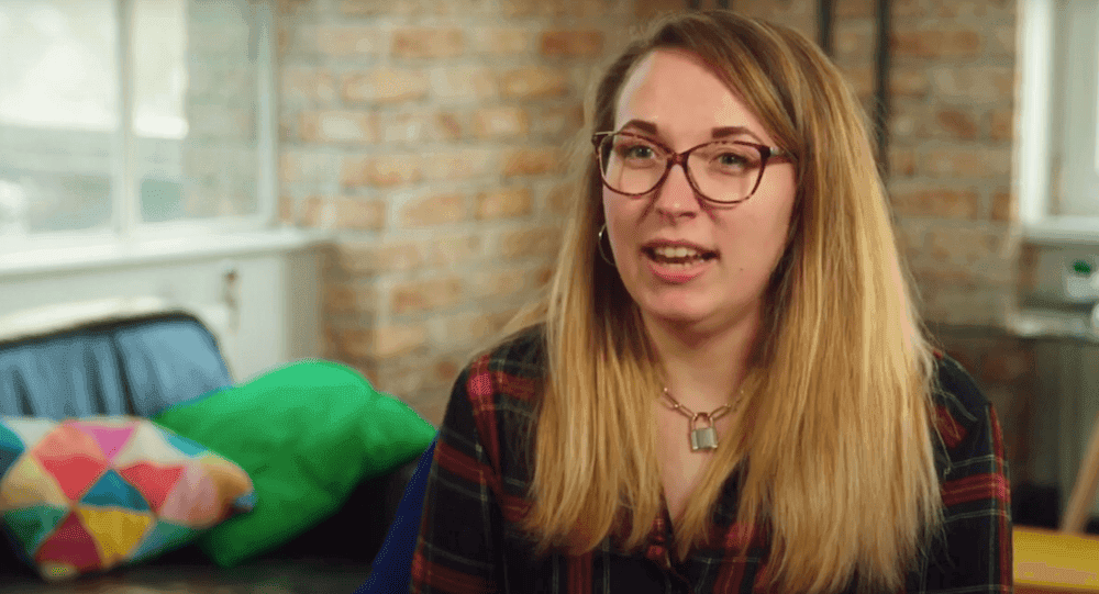 Woman with glasses is wearing a red and black checked shirt sat on a black leather sofa, in front of an exposed brick wall.