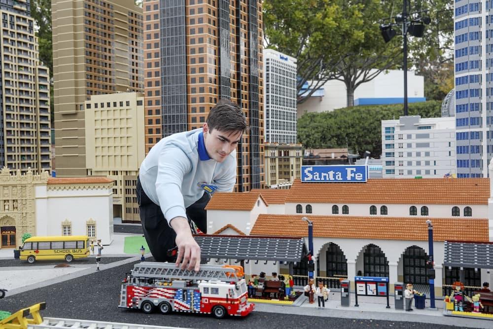 Man is wearing a grey sweatshirt over a blue polo and is rearranging a LEGO® firetruck model in a town made from LEGO® bricks.