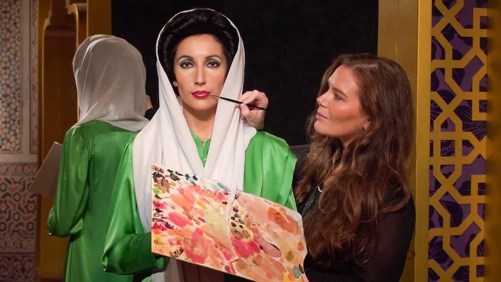Women with long brown hair is holding a paint palette and a paintbrush against the face of a wax model figure.
