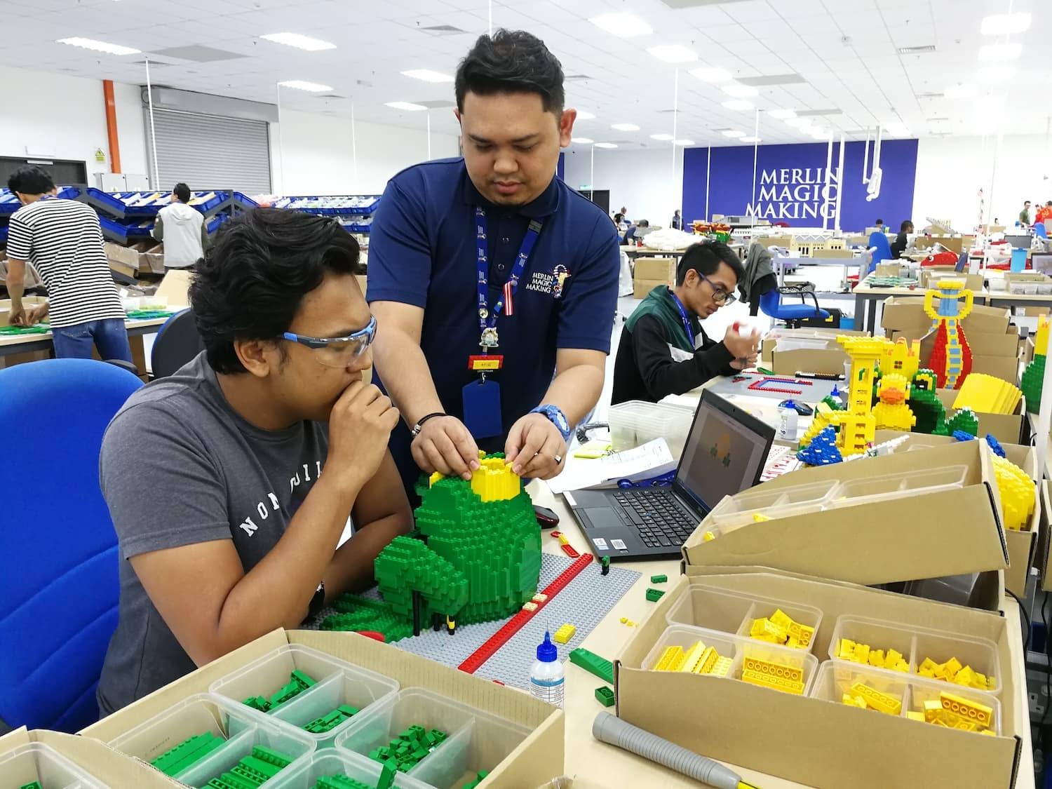 An image of two male LEGO® builders in the Merlin Magic Making area. They are working together to construct a green object made of LEGO® bricks.