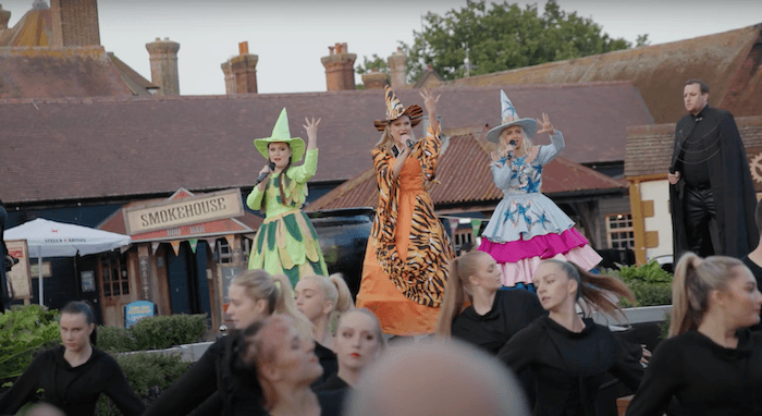 Three women wearing witch dresses and hats are stood on a stage with their hands in the air holding microphones. There is a crowd of entertainers below them.