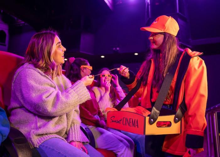 Women wearing orange jacket and cap and concession bucket labelled 'Sunset CINEMA' walking along red chairs.