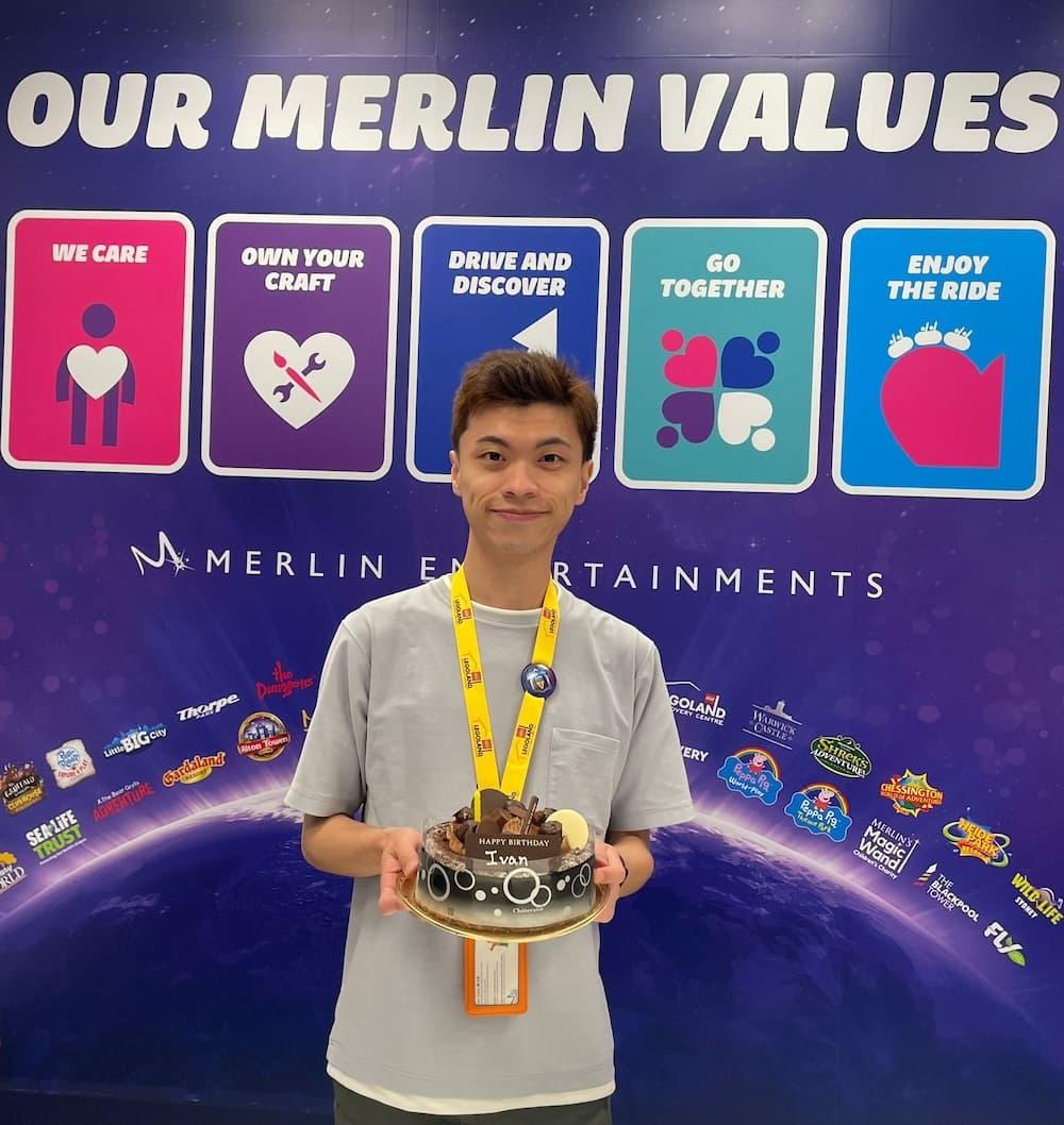 Man is wearing a grey tshirt and a yellow lanyard and is holding a chocolate birthday cake. In the background, there is a display with the text Our Merlin Values with images of Merlin Entertainment logos.